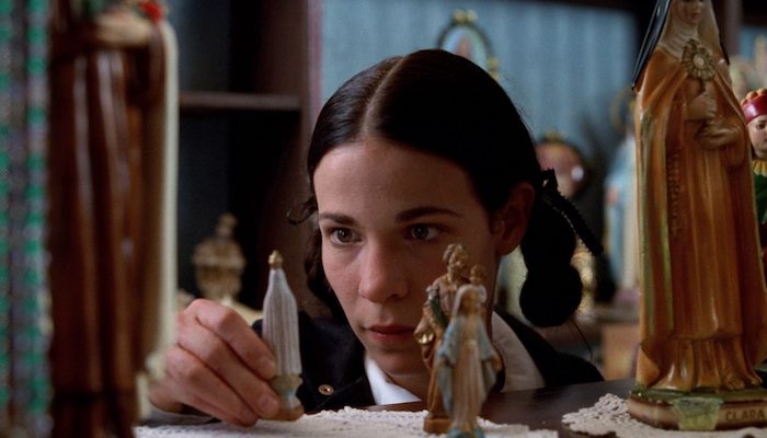 A woman presented in close-up gazes at miniature religious figurines in the foreground.