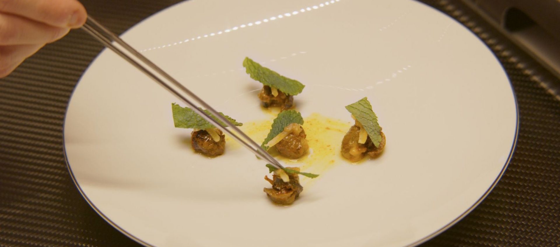 A hand holding chopsticks arranges small bits of food on a white plate. 