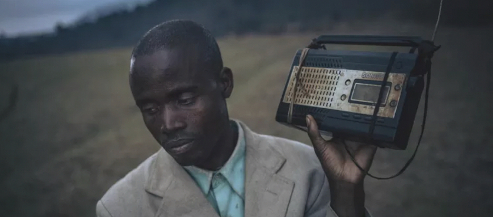 A man with dark skin stands in a field, looking down and holding an old transistor radio close to one ear.