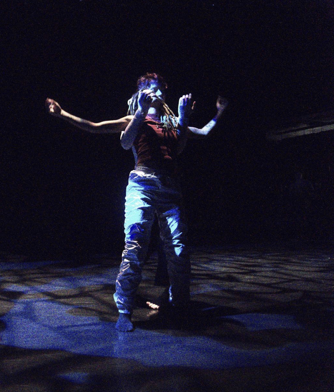 A dancer stands on a dark stage with arms extended while another dancer stands behind them, their arms extended forward under the first dancer’s arms.