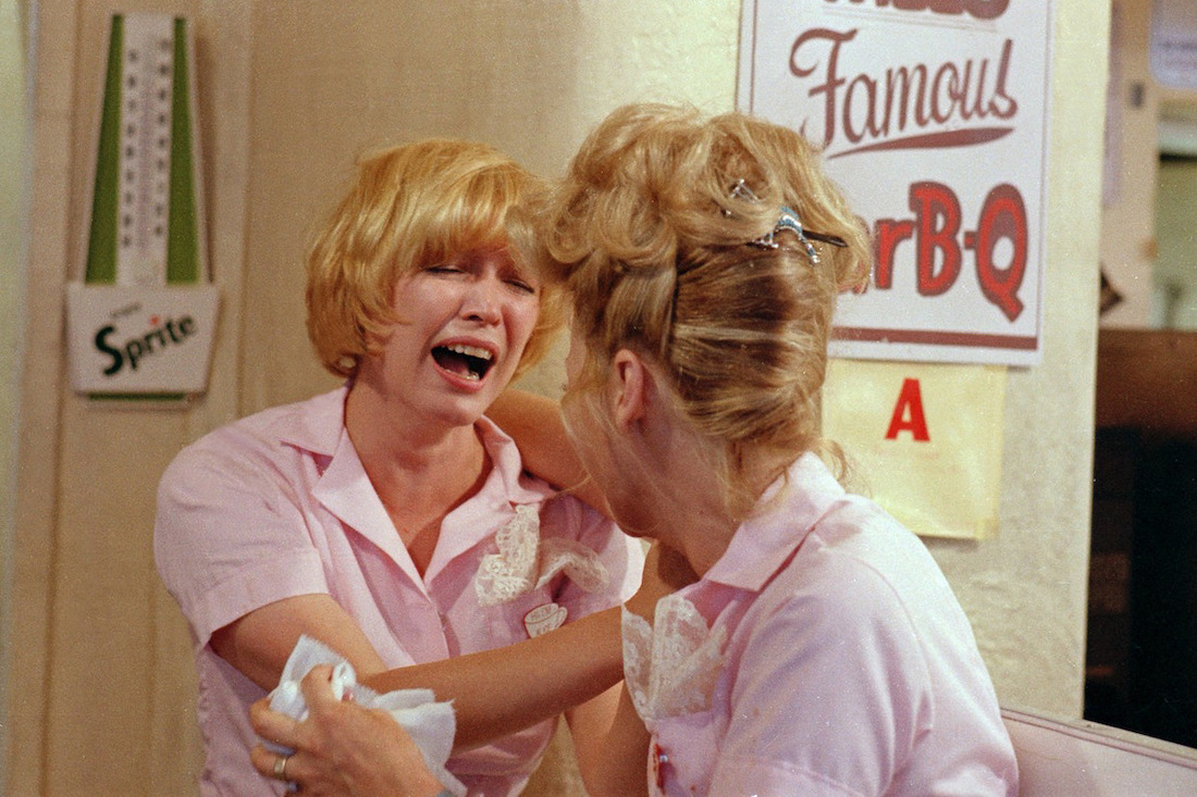 Two uniformed waitresses in a diner face each other as they laugh to the point of tears.