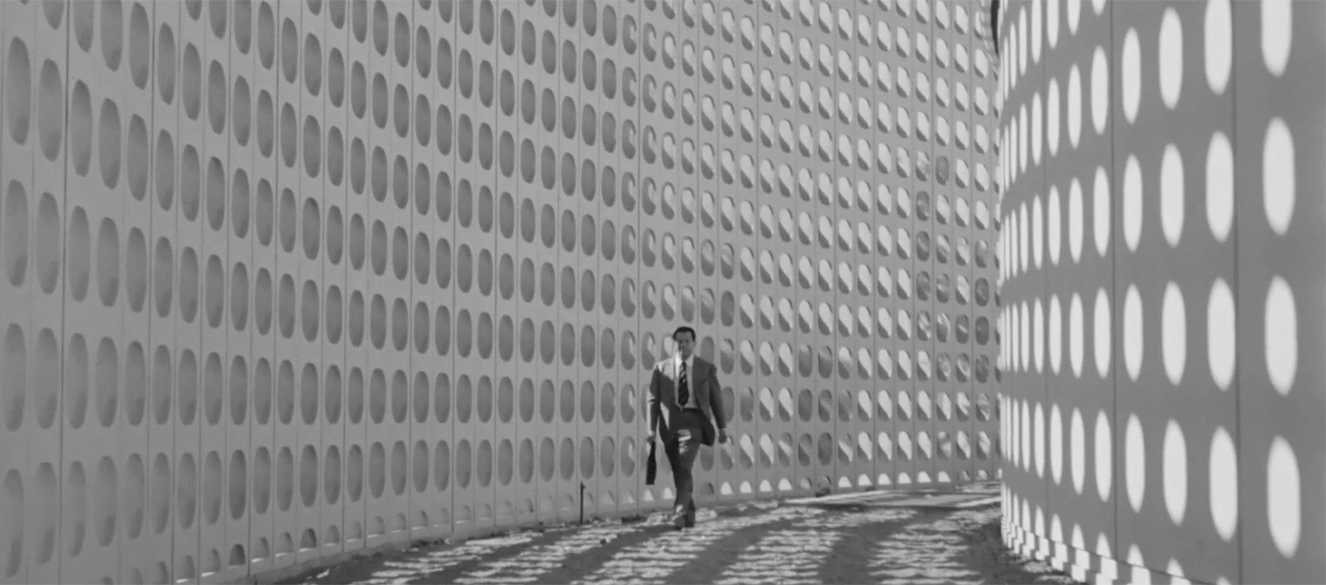A man in a suit is walking in a corridor, one wall has lots of holes that are creating shadows on the opposite wall.