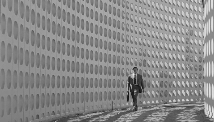 A man in a suit is walking in a corridor, one wall has lots of holes that are creating shadows on the opposite wall.