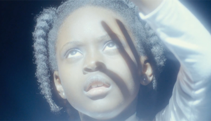 A young Black girl stretches her hand to shield an outpour of light. It casts a shadow against her face.