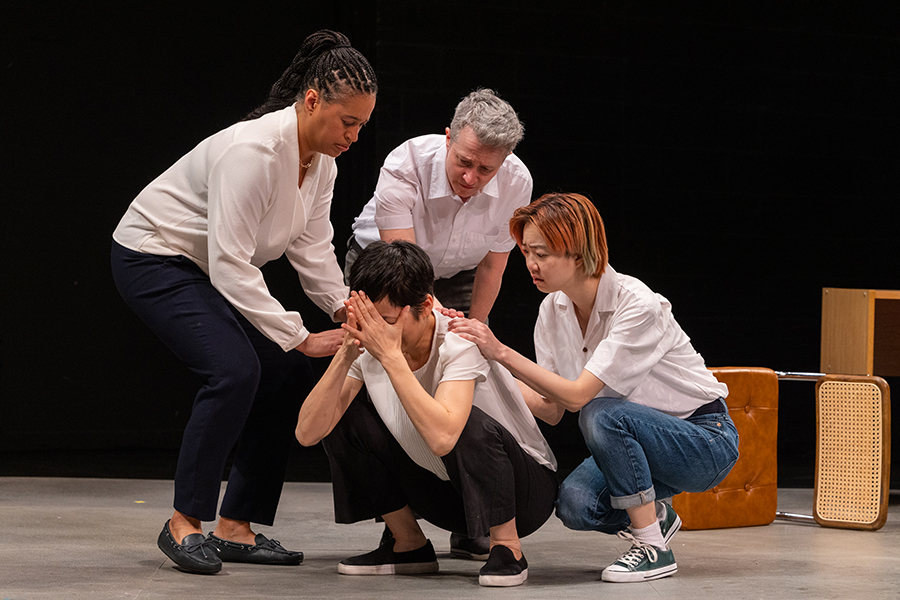 A person with their head in their hands kneels as 3 other actors are consoling them.