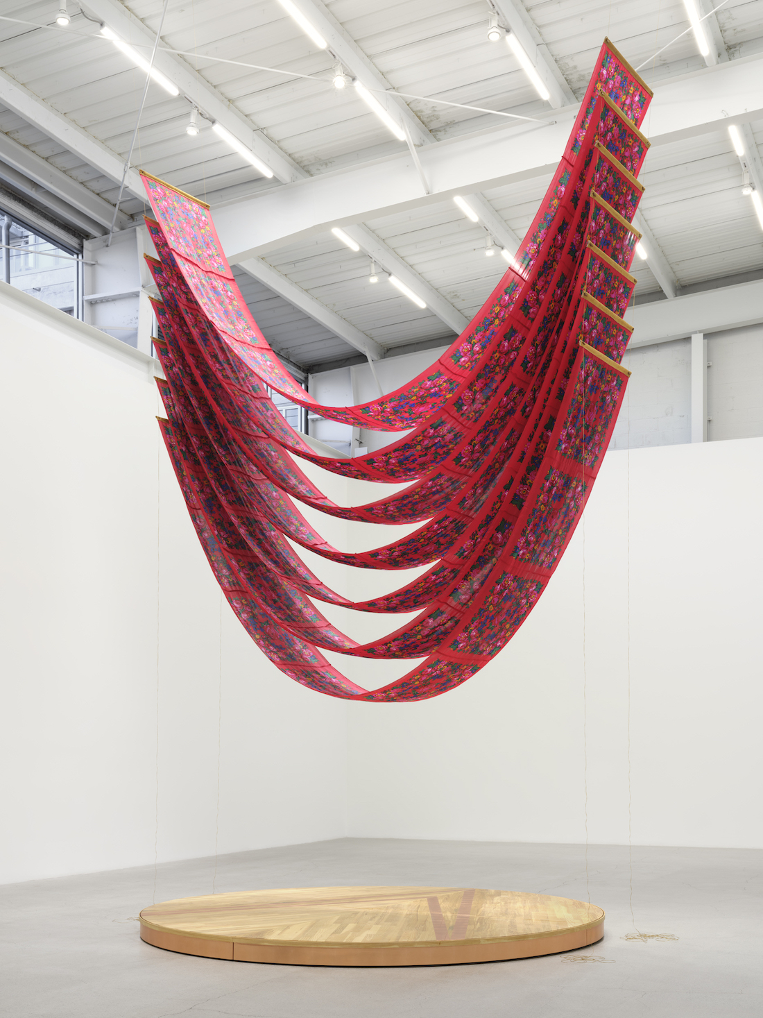 A gallery installation of long, narrow bands of printed red fabric hung by both ends in a dangling u-shape over a round wooden platform.