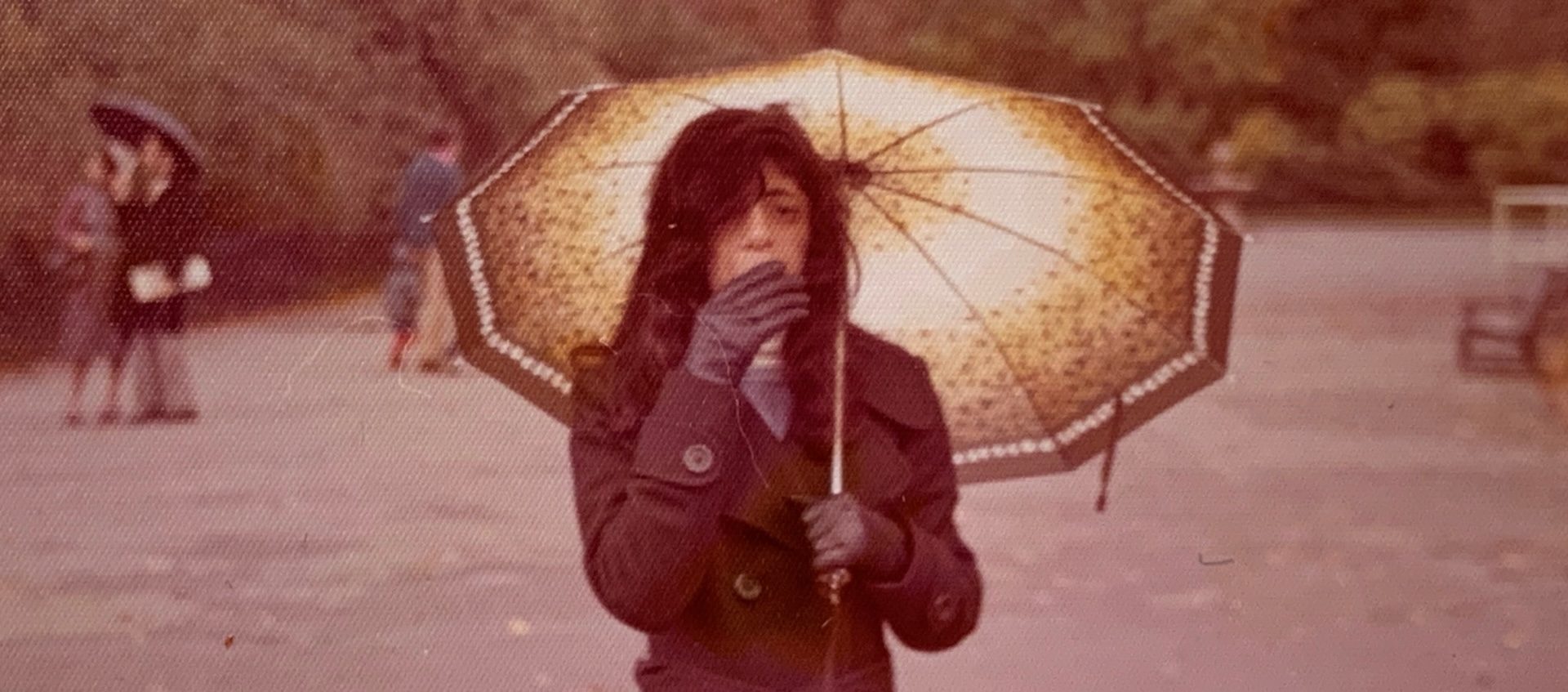 A person with long dark hair stands outdoors under an umbrella. Their gloved hand partially covers their face.