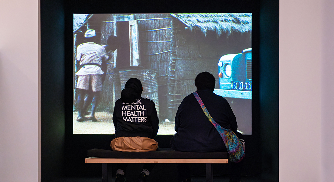 Two people sit in a small room facing a film screen. One wears a sweat shirt that reads Black Mental Health Matters and the other has colorful bag at their side and wears a dark shirt.