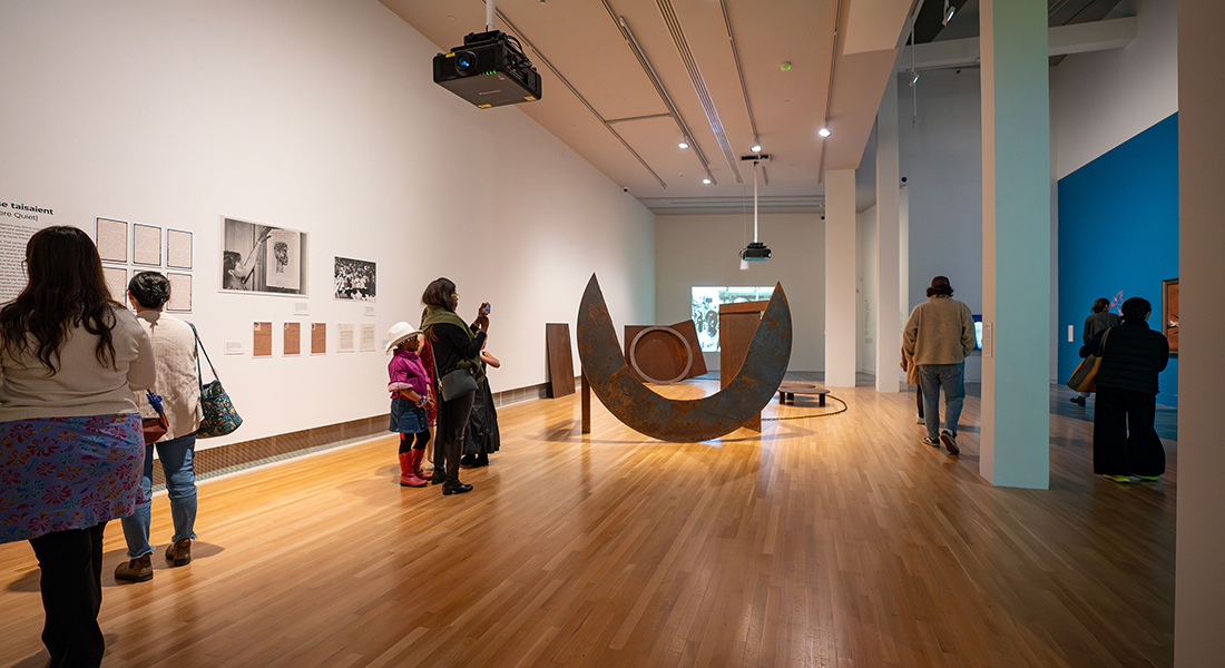 A wide view of a contemporary gallery space. It holds several people looking at various wall artworks or a large metal sculpture at the center of the space.