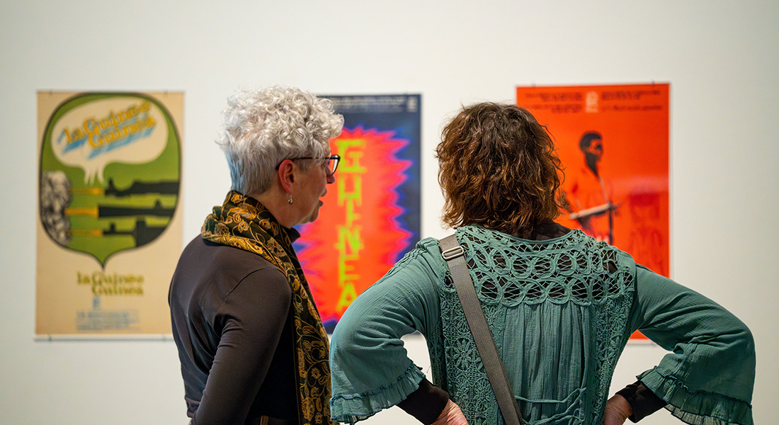 Two people are captured from behind as they look at a row of revolutionary posters on a gallery wall.
