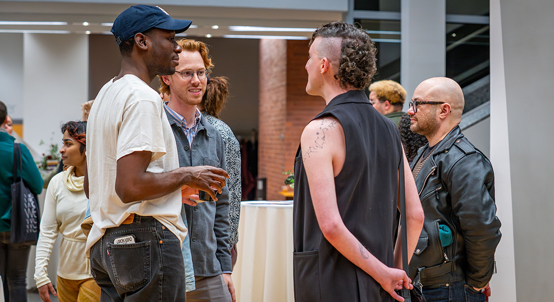 Three people stand together during an exhibition opening in the Wexner Center lower lobby and talk, as others stand around them.