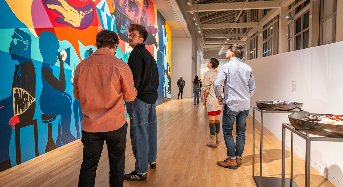 Several people stand in a contemporary gallery space and face a large, colorful wall mural.