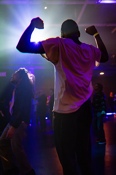 A person is captured from behind as they dance during a DJ set. They're silhouetted by an overhead light in front of them.