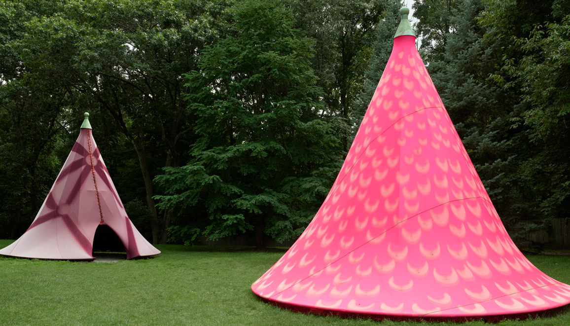  Two pink teepee-shaped structures, each with different pink patterned cloth, sit in a grassy field with trees in the background.