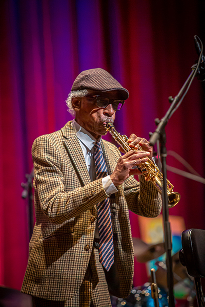 Roscoe Mitchell plays the soprano saxophone on a red and violet lit stage. He wears a brown plaid jacket, glasses, and brown flat cap.