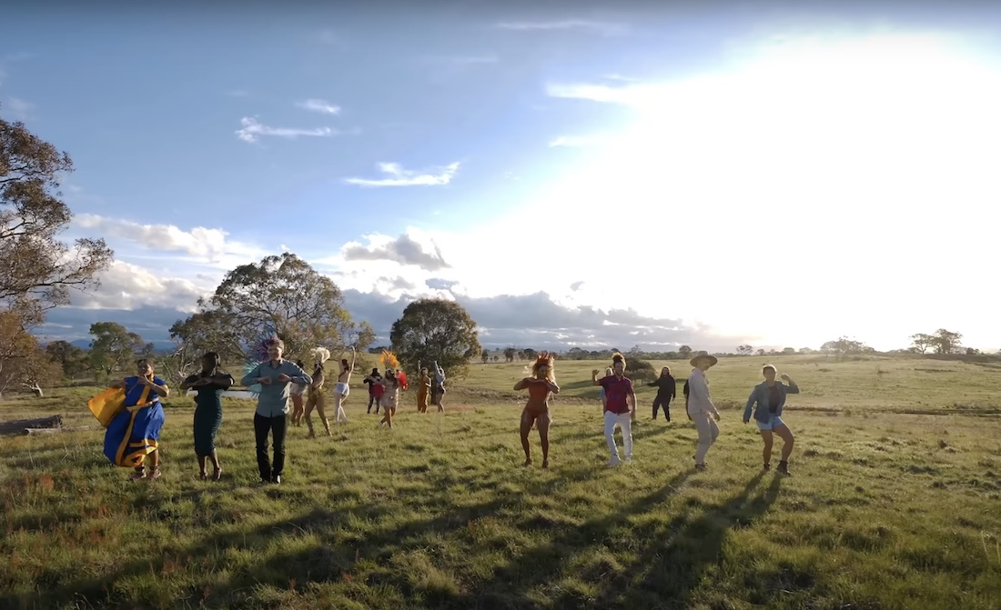 A group of over a dozen people in various costumes or street clothes dance outside on a wide flatland under a bright sun.