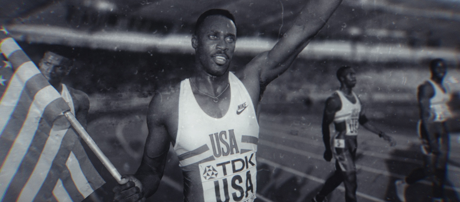 A Black man stands on a race track. He holds an American flag with one hand and raises his other arm in victory.