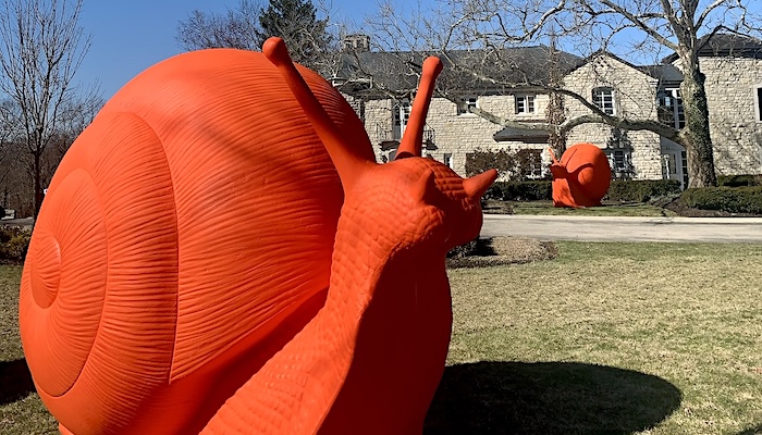 Two large orange snail sculptures sit outside in front of a large stone building.