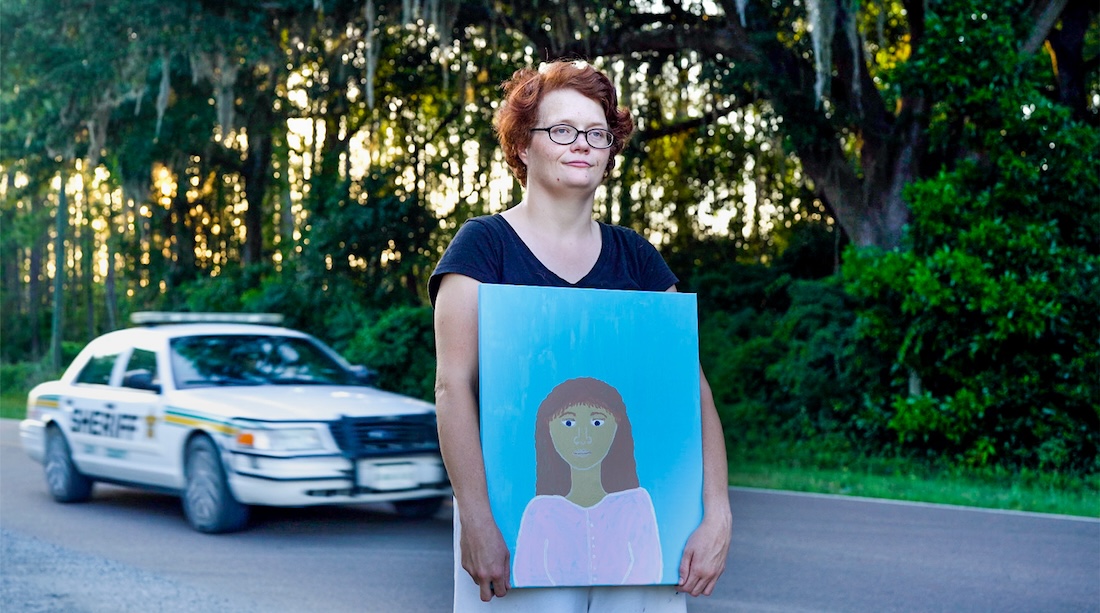 A woman stands in front of a police car, looking forward and holding a painting of a girl.