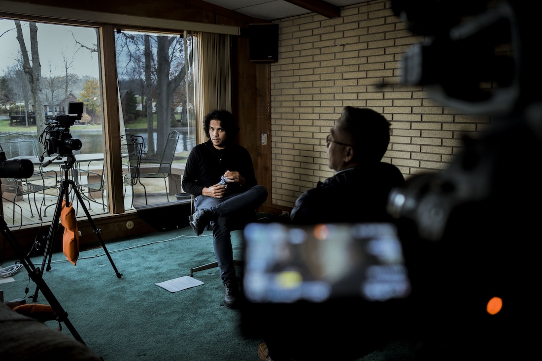 A young man of Filipino descent sits surrounded by cameras, preparing to interview someone.