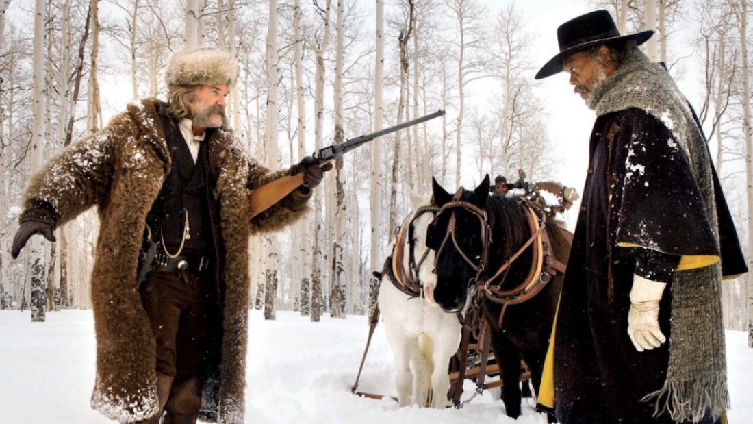 A still from a movie Western of one man pointing a rifle at close range at another man as their horses look on. They stand in a remote, snow-covered forest.