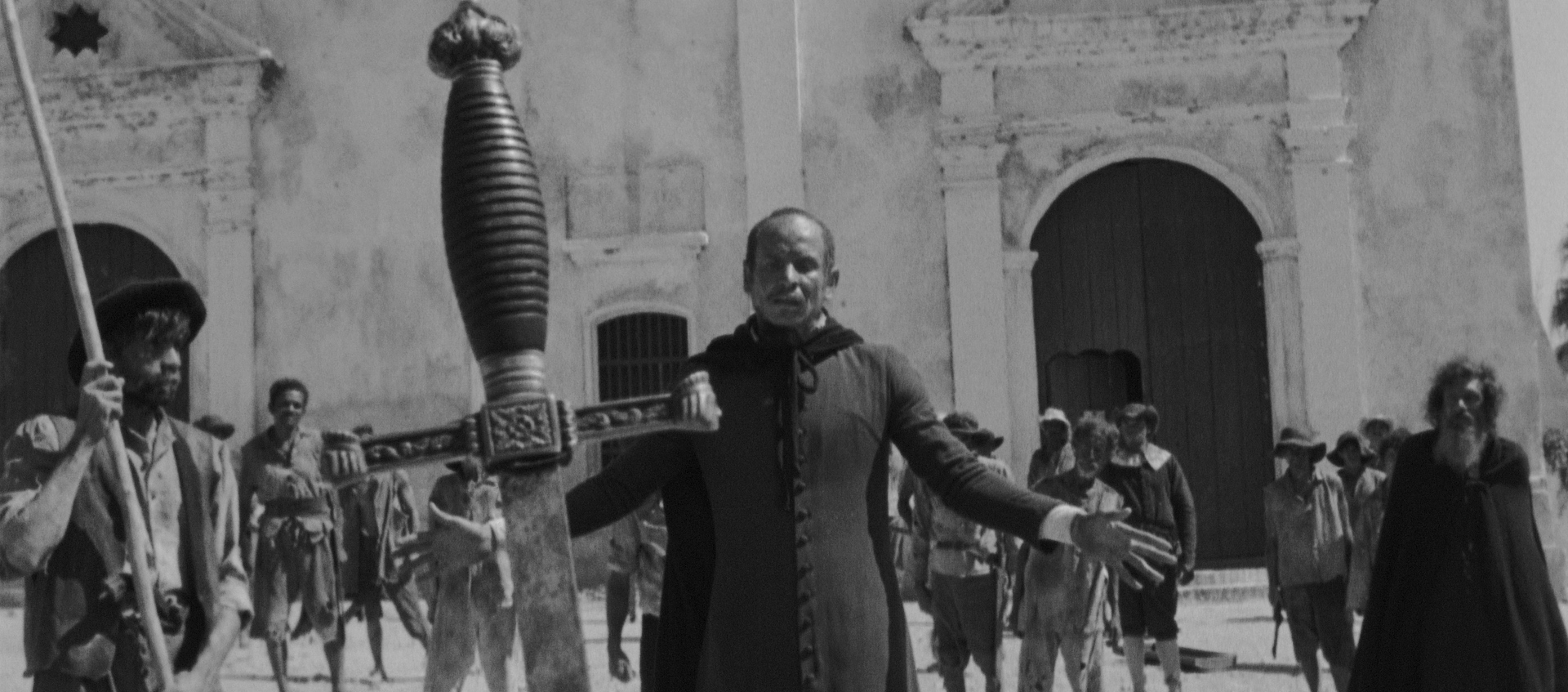 A man in religious garb stands with his hands out to his sides in front of a large stone building behind a giant sword stuck in the ground.