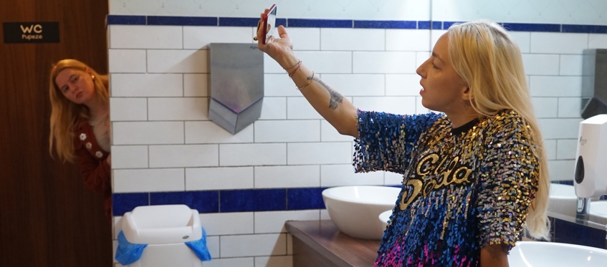 A blond woman in a colorful, sequined shirt takes selfies in a large, clean bathroom while another woman peers her head into the open doorway.