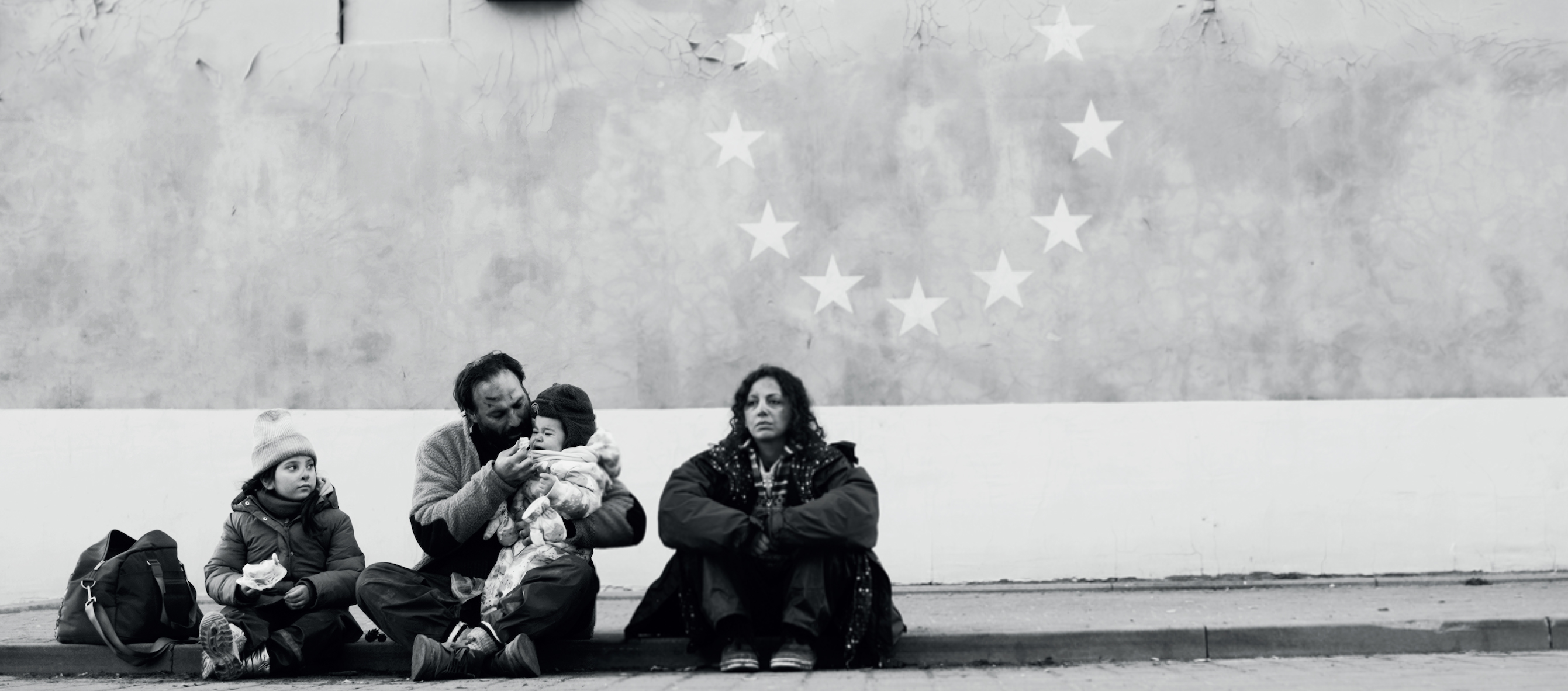 A family of four sits on the ground wearing winter coats in front of a wall with a circle of stars on it.