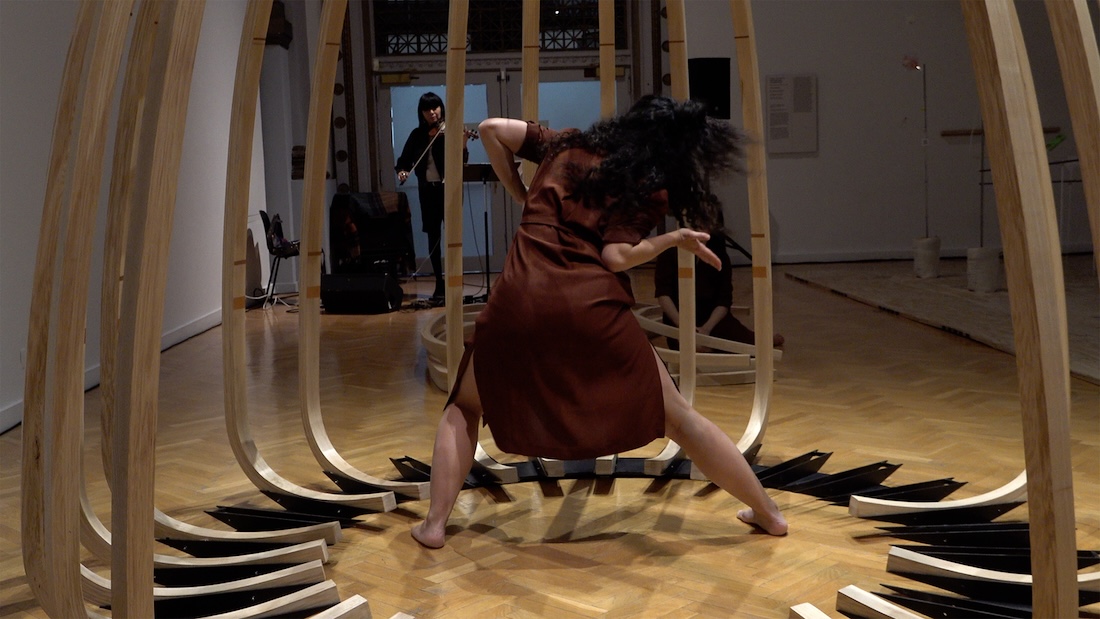 A woman is seen from behind, dancing within a whale bone-line sculptural structure, while a violinist plays in the background.