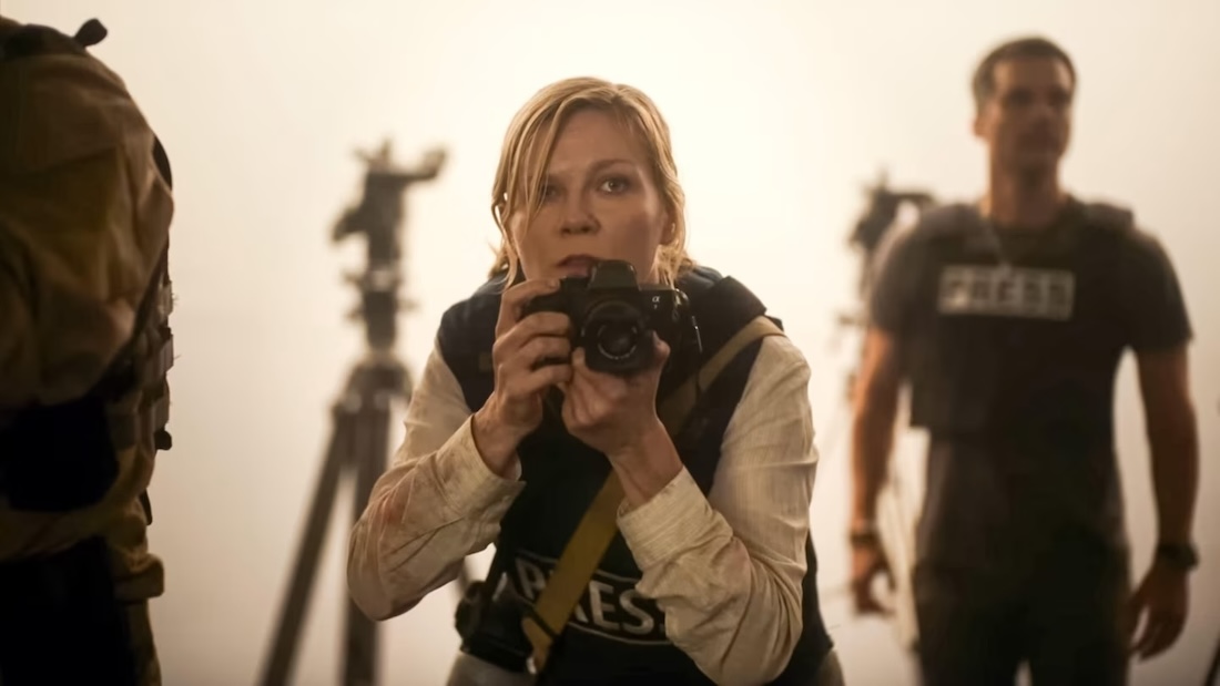 A woman wearing a press vest holds a camera close to her face as she gazes with fear at something in front of her.