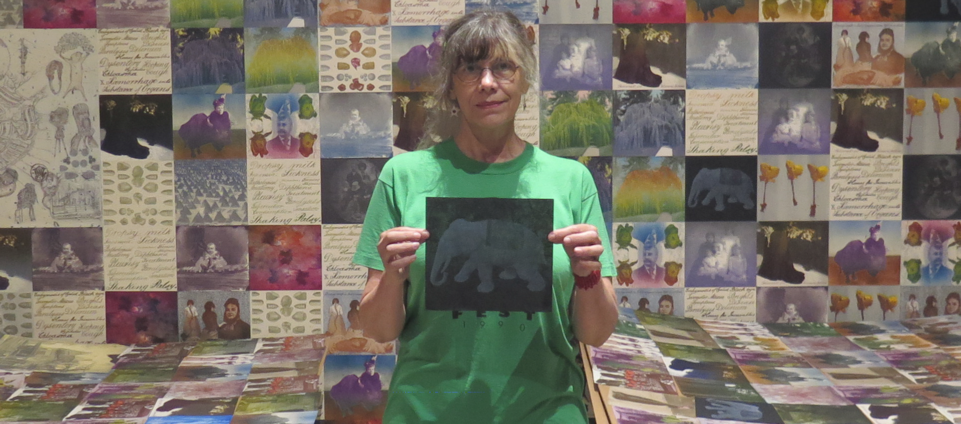 A woman with graying hair and glasses stands in front of a wall papered with square artworks and holds up a pictures of an ivory elephant sculpture.