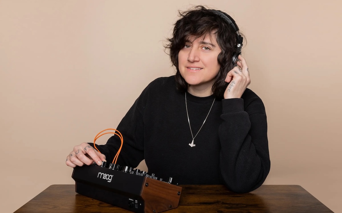 A person with short dark hair and a slight smile sits at a wooden table wearing headphones attached to a Moog synthesizer. 