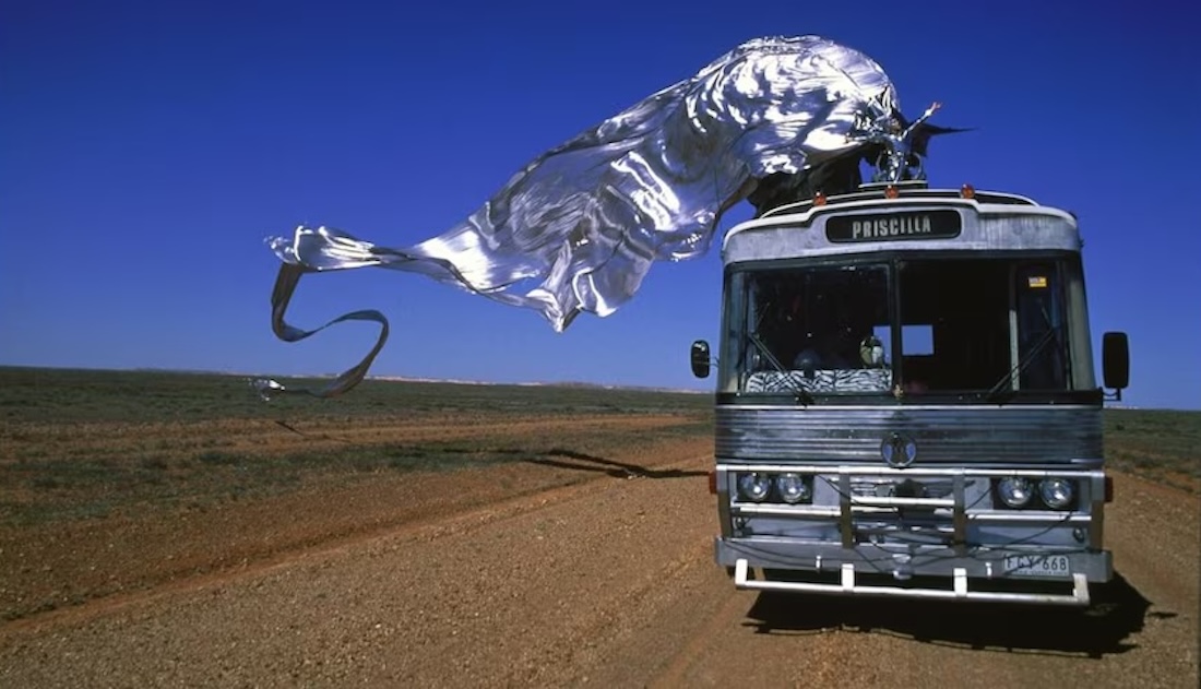 A vintage bus travels on a road in the Australian outback while a drag queen in a billowing silver cape performs on the bus’ roof.