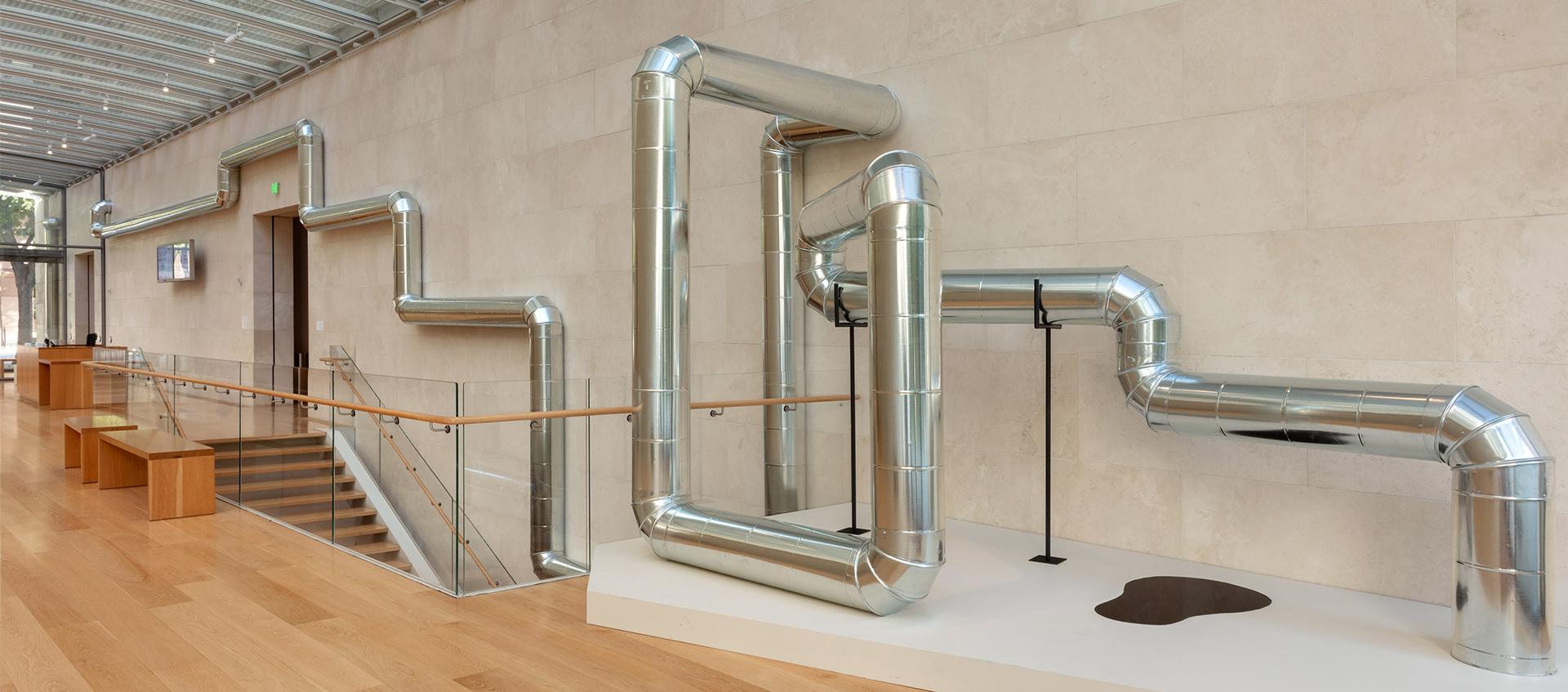 An industrial steel pipeline hangs on a wall above a museum stairwell. It frames architectural elements and ends on a plinth with a pool of oil on it.