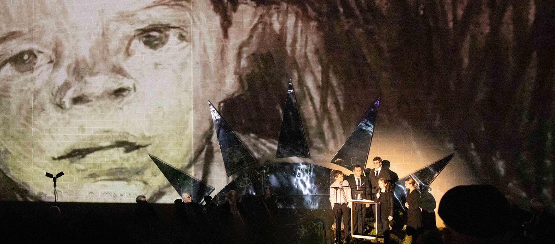 An image from the original production of Calling Hours, a large drawing of a child’s face is projected behind a group wearing suits and black dresses. 