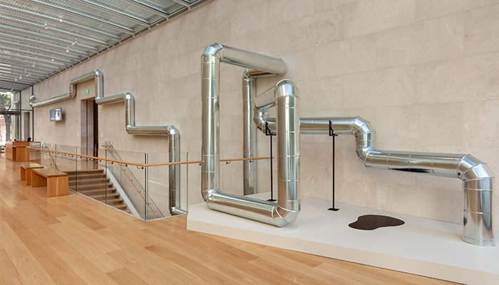 An industrial steel pipeline hangs on a wall above a museum stairwell. It frames architectural elements and ends on a plinth with a pool of oil on it.
