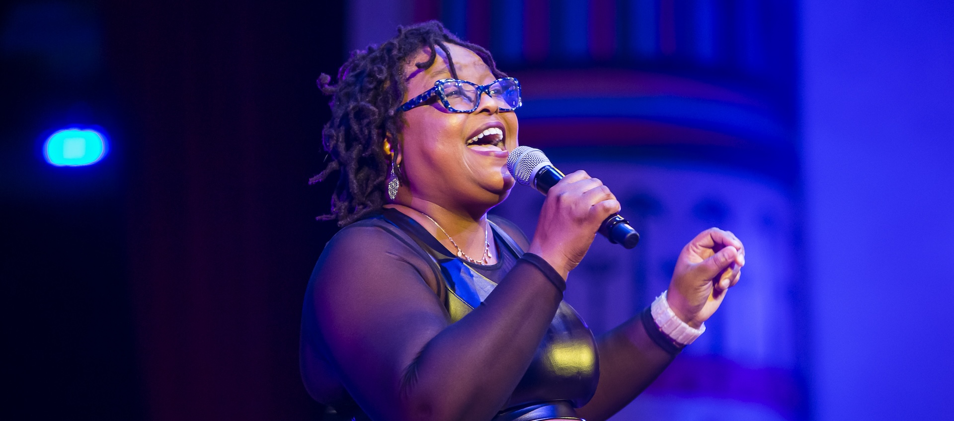 A Black woman with short, tight dreadlocks and glasses smiles as she stands on a stage and sings into a microphone.