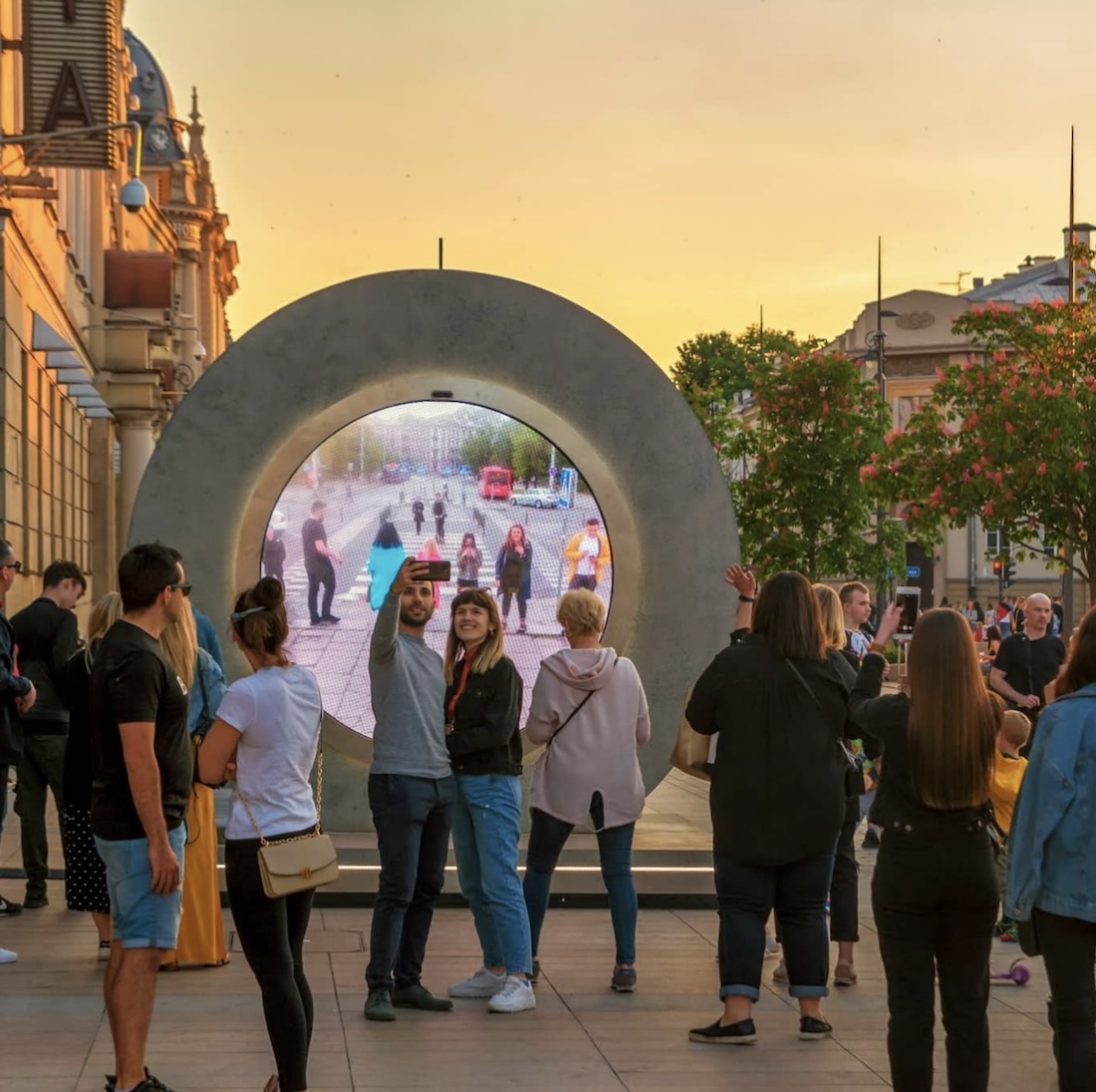 A small crowd of people gather around a circular sculpture with a link to video from a different street in its center.