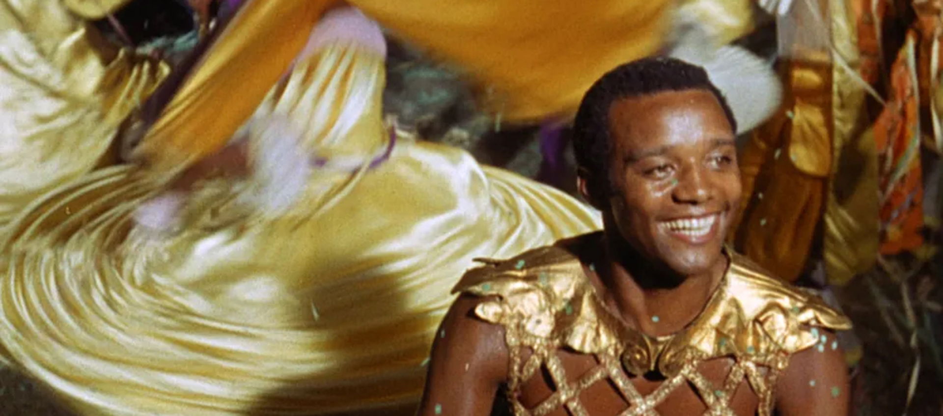 A young Black man wearing Roman garb sits in front of twirling dancers.