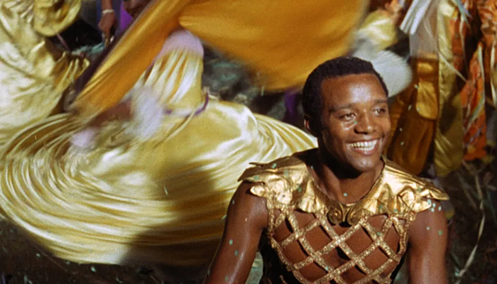A young Black man wearing Roman garb sits in front of twirling dancers.