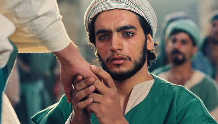 A man with facial hair wearing a headcovering kneels while holding someone's hand.
