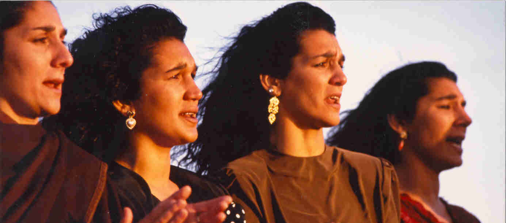 Four women stand together with their hair blowing in the wind. They appear to be singing.