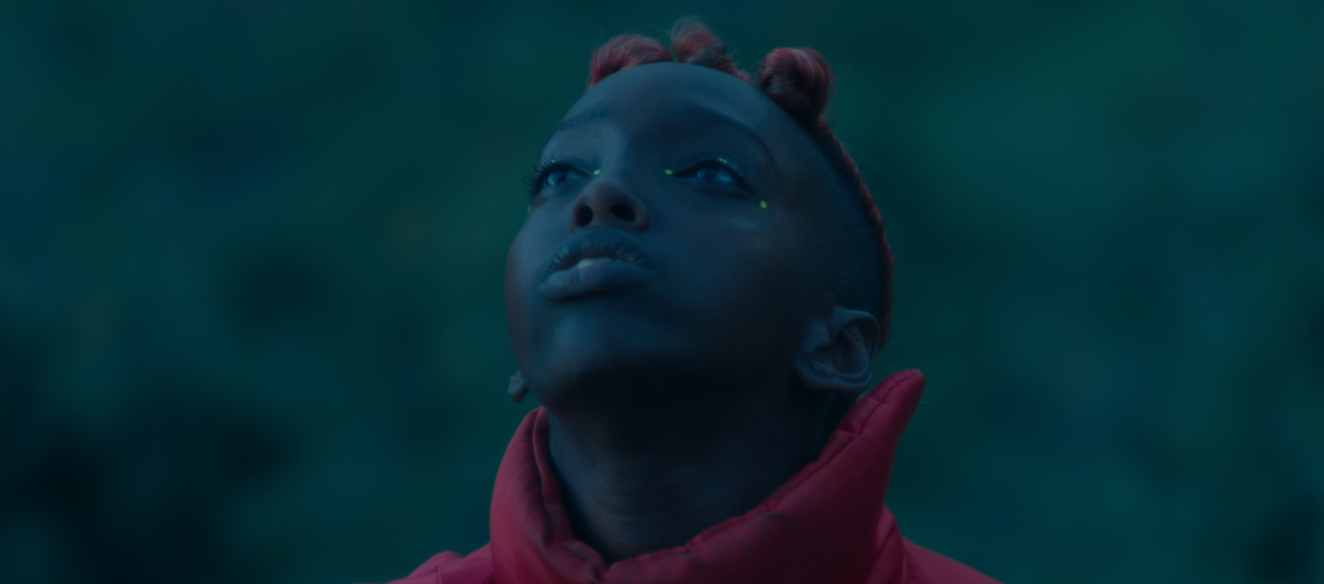 A boy looks up he is wearing a red shirt with a high collar and has braids and dots on either side of his eyes.
