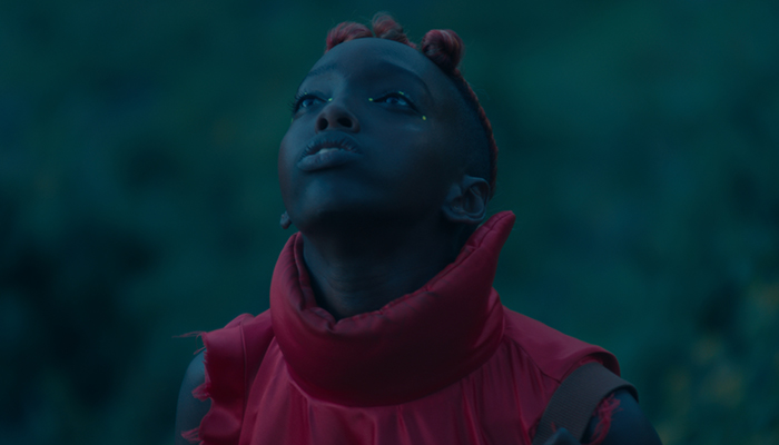 A boy looks up he is wearing a red shirt with a high collar and has braids and dots on either side of his eyes.