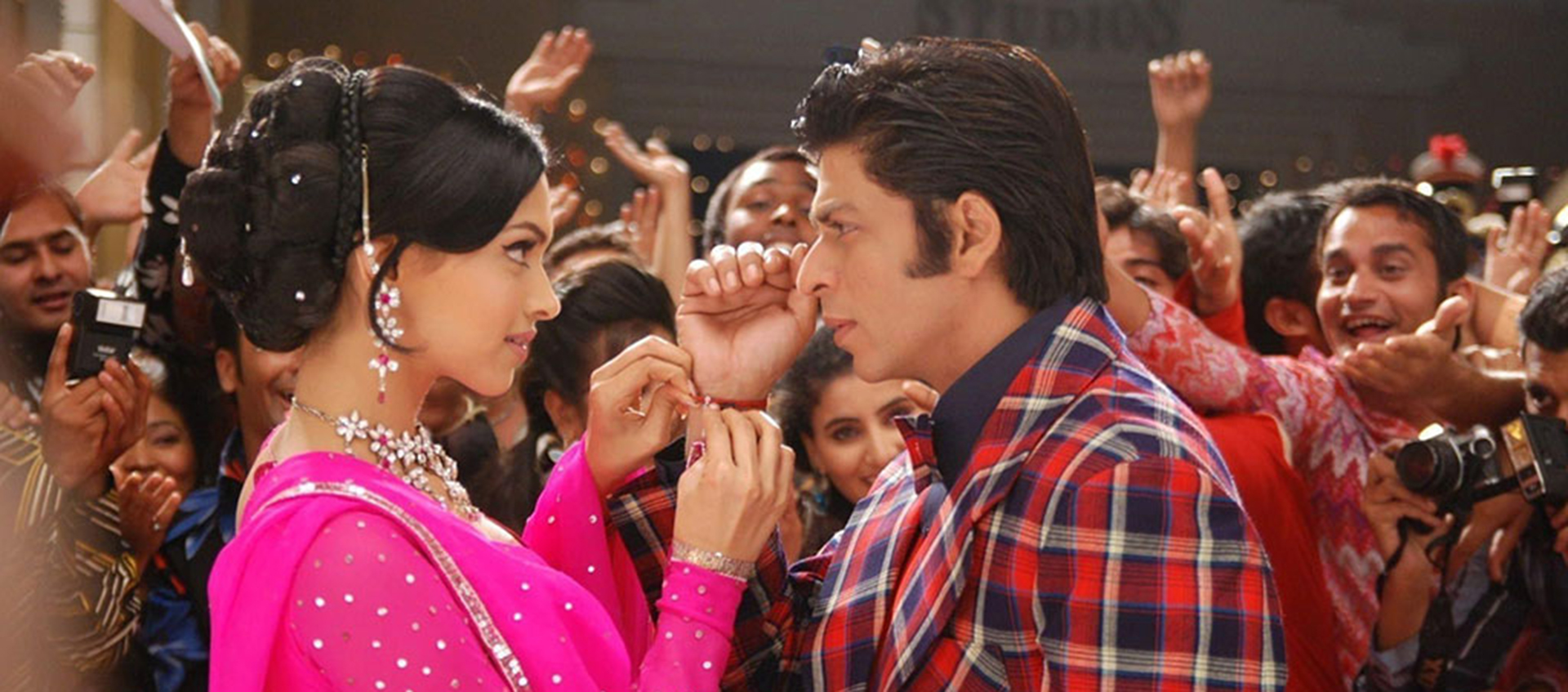 A man and woman stand facing eachother surrounded by a crowd. She wears a hot pink sari and he wears a plaid suit.