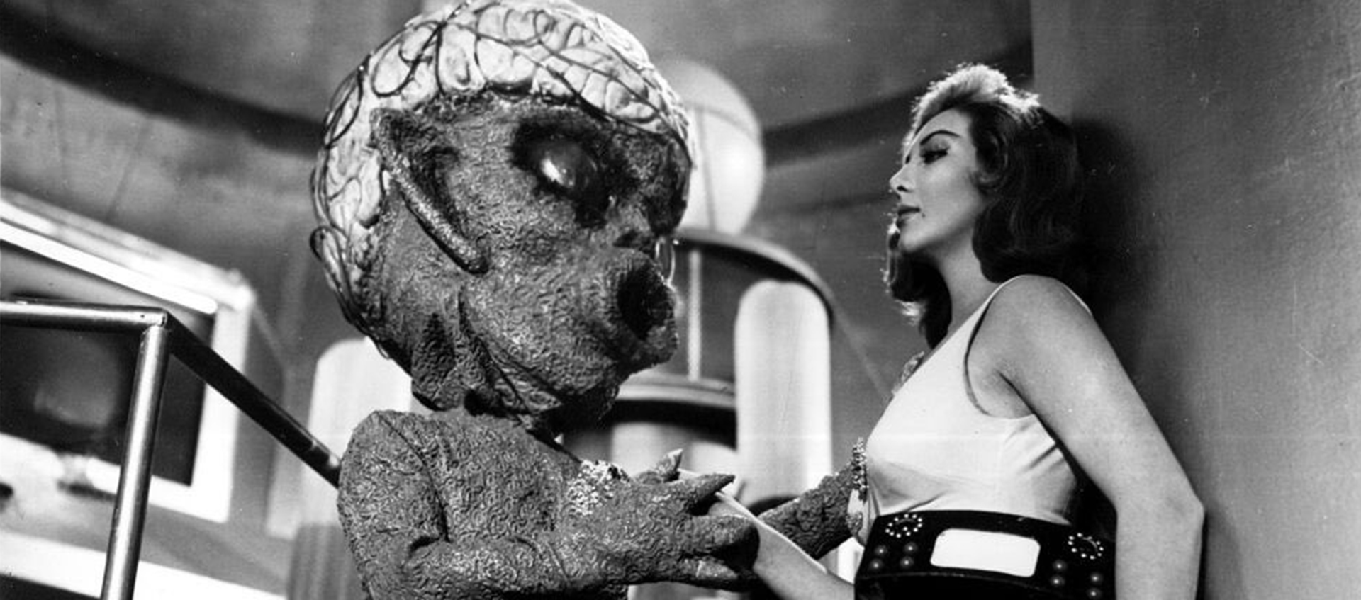 An alien with a large head holds a woman against a wall.
