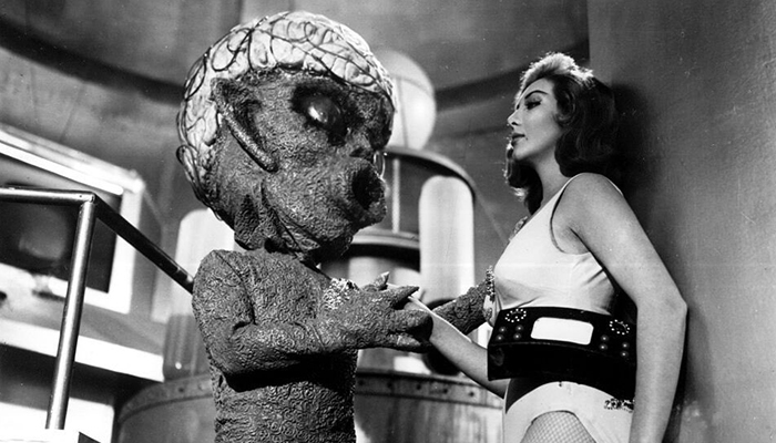 An alien with a large head holds a woman against a wall.