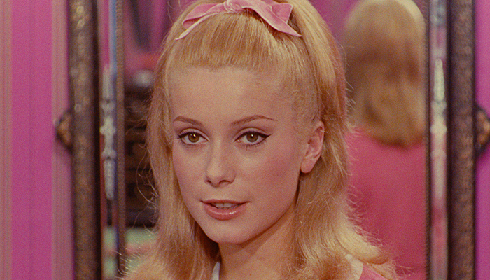 A young blond woman faces away from a mirror hanging on a pink wall.