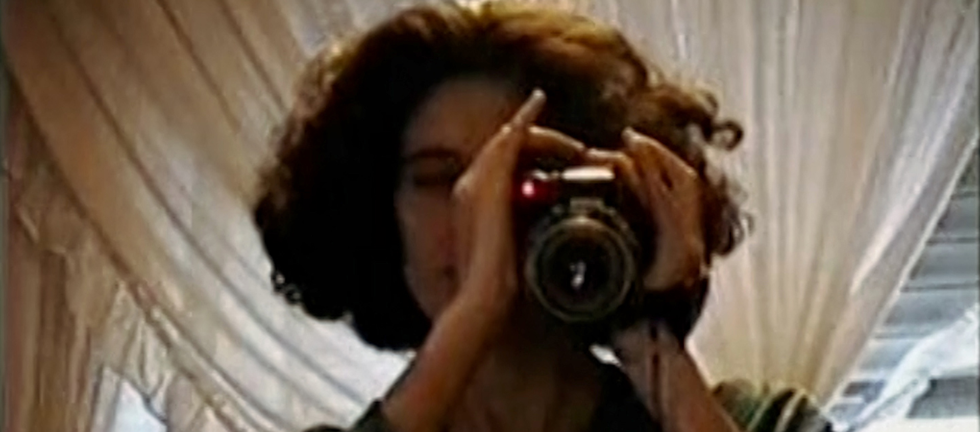 a woman with short curly hair holds a camera up to her face.