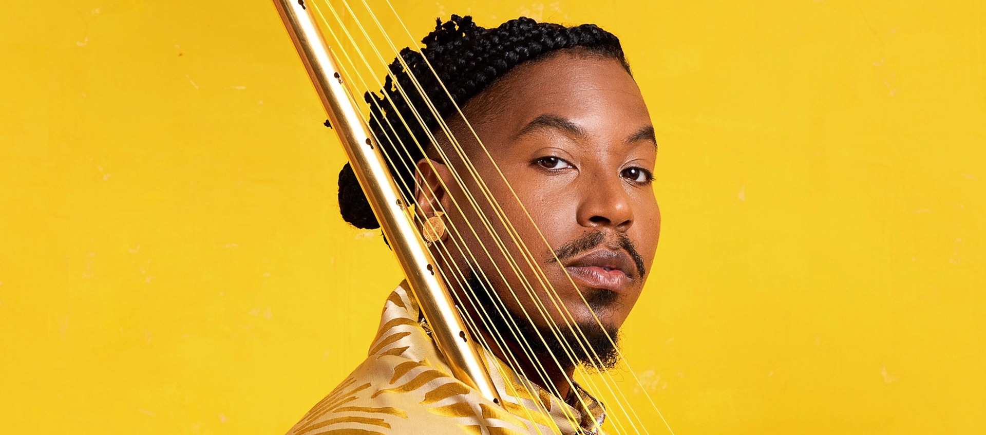 Headshot of Chief Adjuah. He wears a gold shirt against a yellow background and holds a stringed instrument. 
